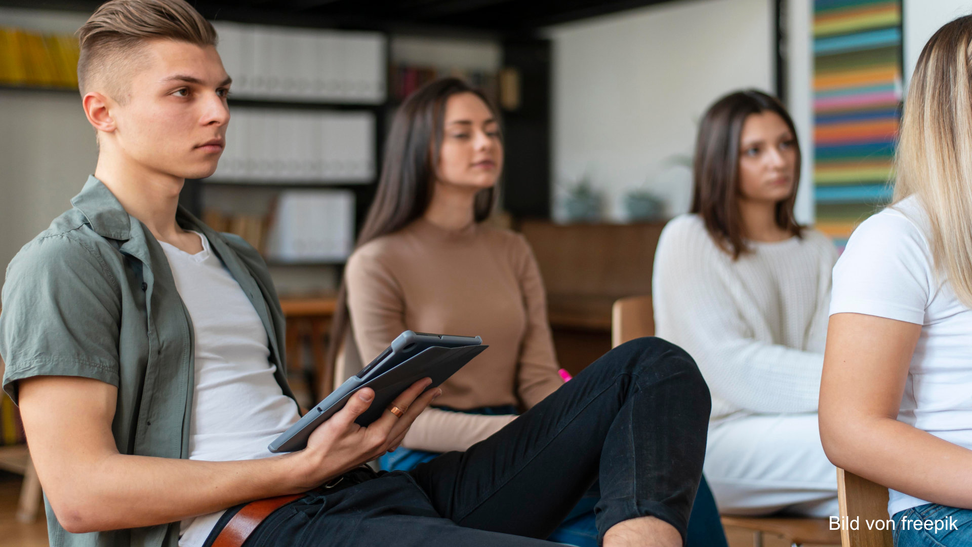 Young people sitting in a classrom. Focus on a young men with a tablet in hand, listening to a speaker.